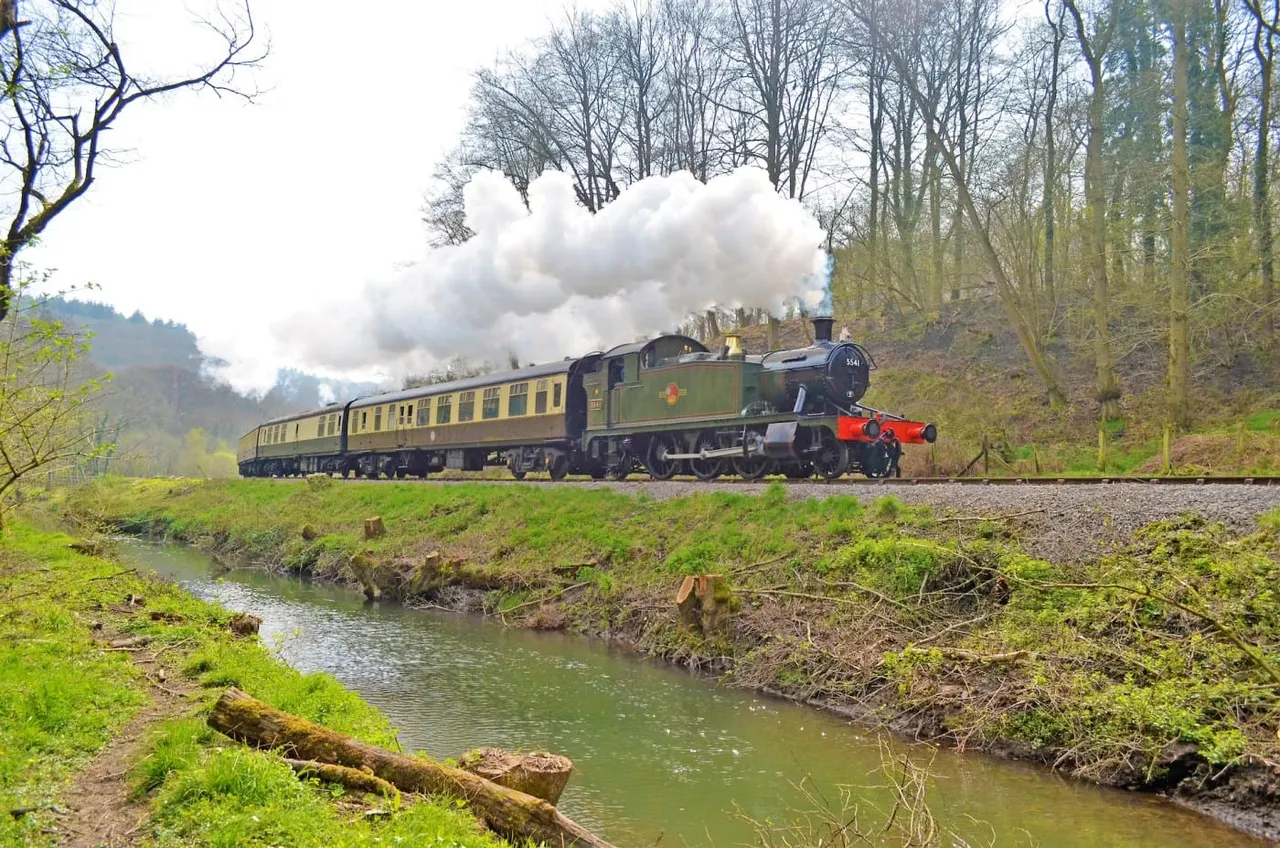 Steam locomotive with yellow passenger carriages travelling through a wooded valley alongside a stream.