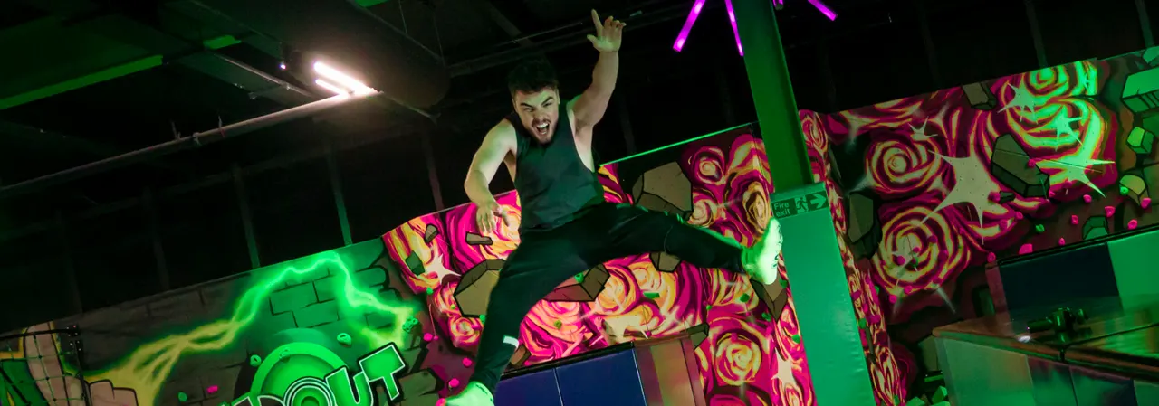 Person jumping on a brightly lit trampoline park with neon green and pink floral-patterned walls and neon tubing overhead.