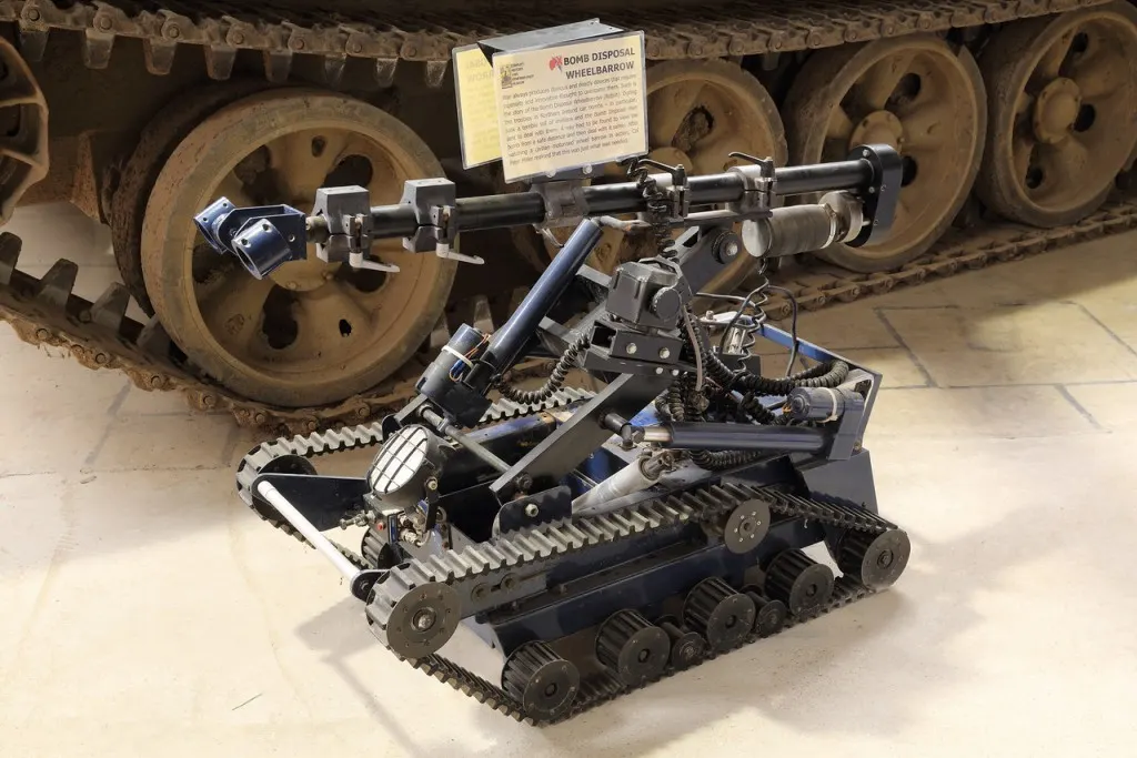A bomb disposal wheelbarrow robot displayed indoors with tracked treads and robotic arm, positioned in front of a large military tank.