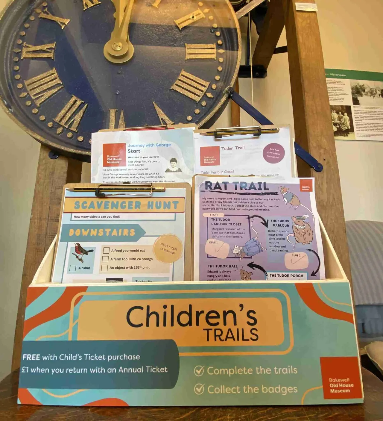 Children's trail activity leaflets and maps displayed beneath a large antique clock with Roman numerals in a museum setting.