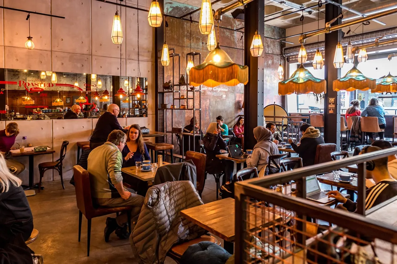 Busy modern café interior with customers seated at wooden tables beneath hanging pendant lights and decorative umbrellas, industrial brick walls and steel beams visible.