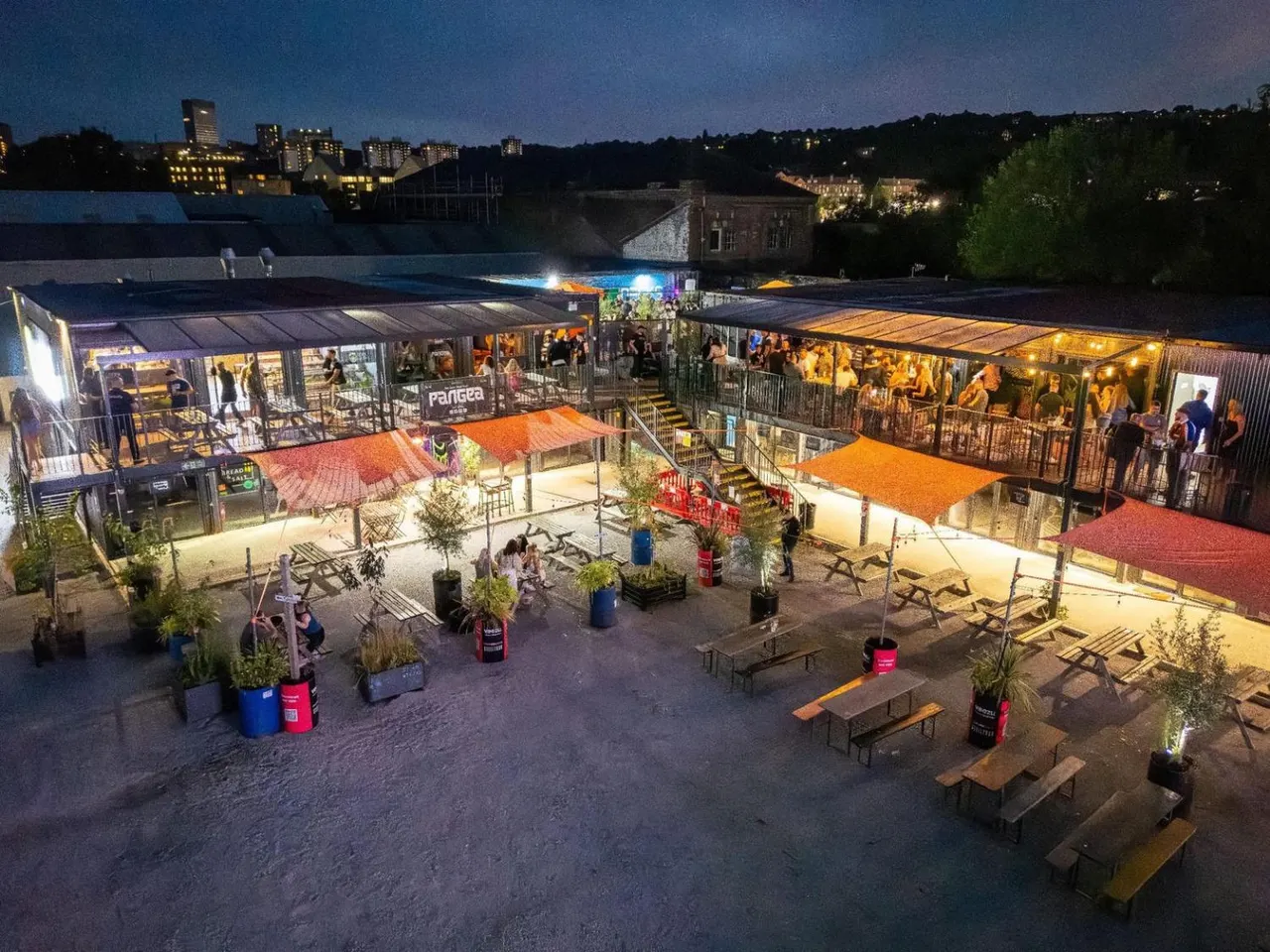 Illuminated outdoor venue at dusk with food stalls, colourful canopies, seating areas and visitors gathered on a sandy ground beside a waterway.