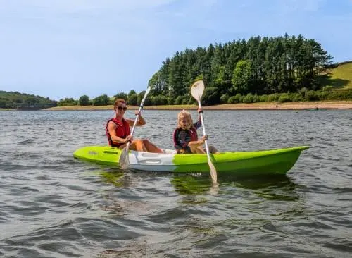 Two children paddling a green kayak on a lake with forested hills in the background.