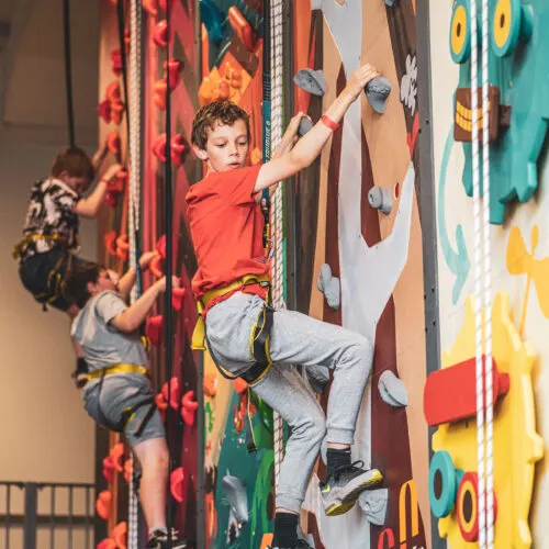 Child in red shirt climbing an indoor climbing wall with coloured holds while wearing a safety harness.