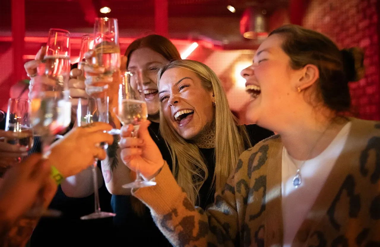 Group of friends laughing and toasting with drinks at a bar with red ambient lighting.