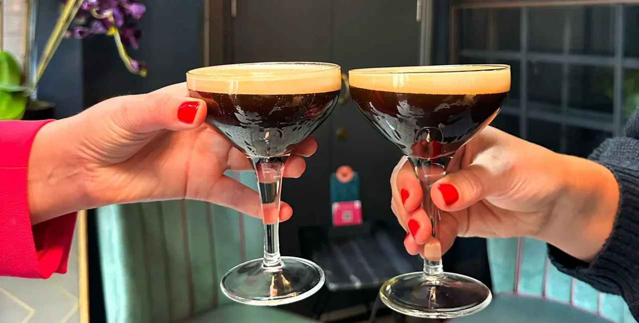 Two people with red nail polish toast with espresso martinis topped with creamy foam.