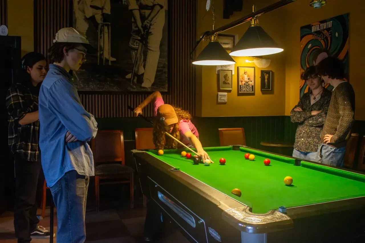 Young people playing snooker at a pool table in a warm-lit pub with vintage decor and hanging lamps.