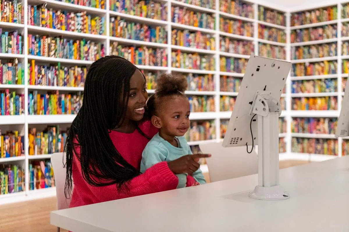 Woman and young child using a computer terminal in a library with colourful bookshelves behind them.