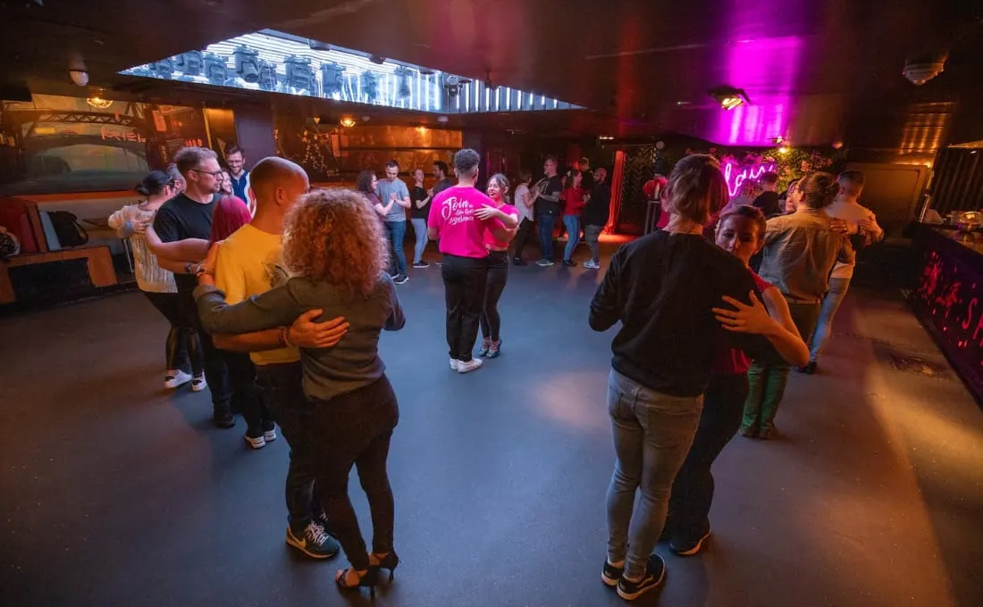 Groups of people dancing together in a colourful lit indoor venue with neon signs and a digital display overhead.