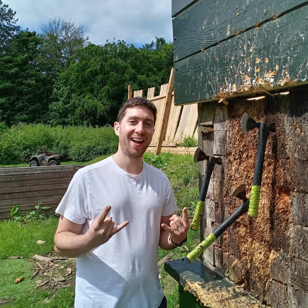 Man smiling at camera whilst holding axe next to wooden structure at outdoor activity venue.