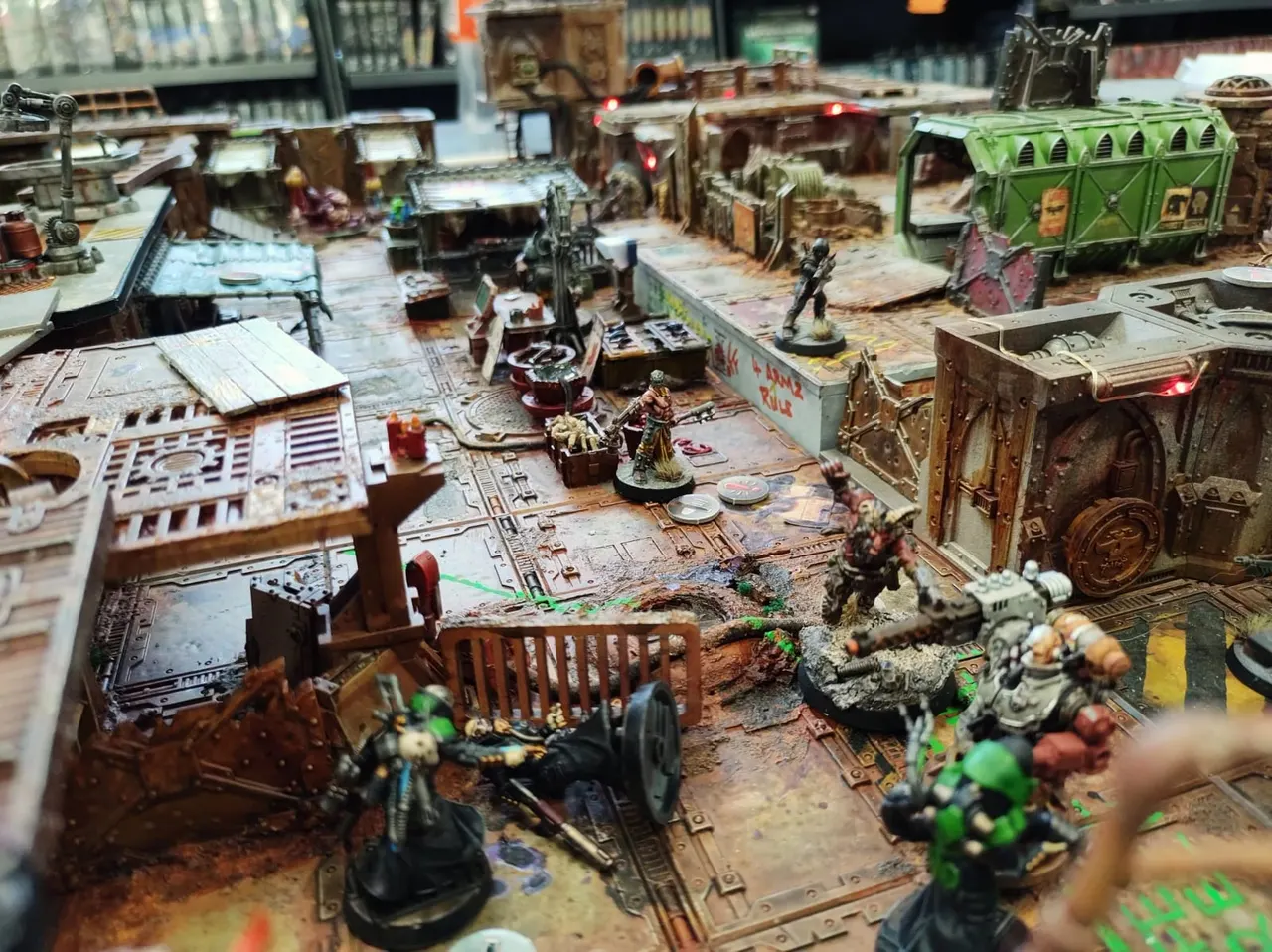 Detailed tabletop wargaming scene with painted miniature figures, buildings, and vehicles arranged on a textured gaming board.