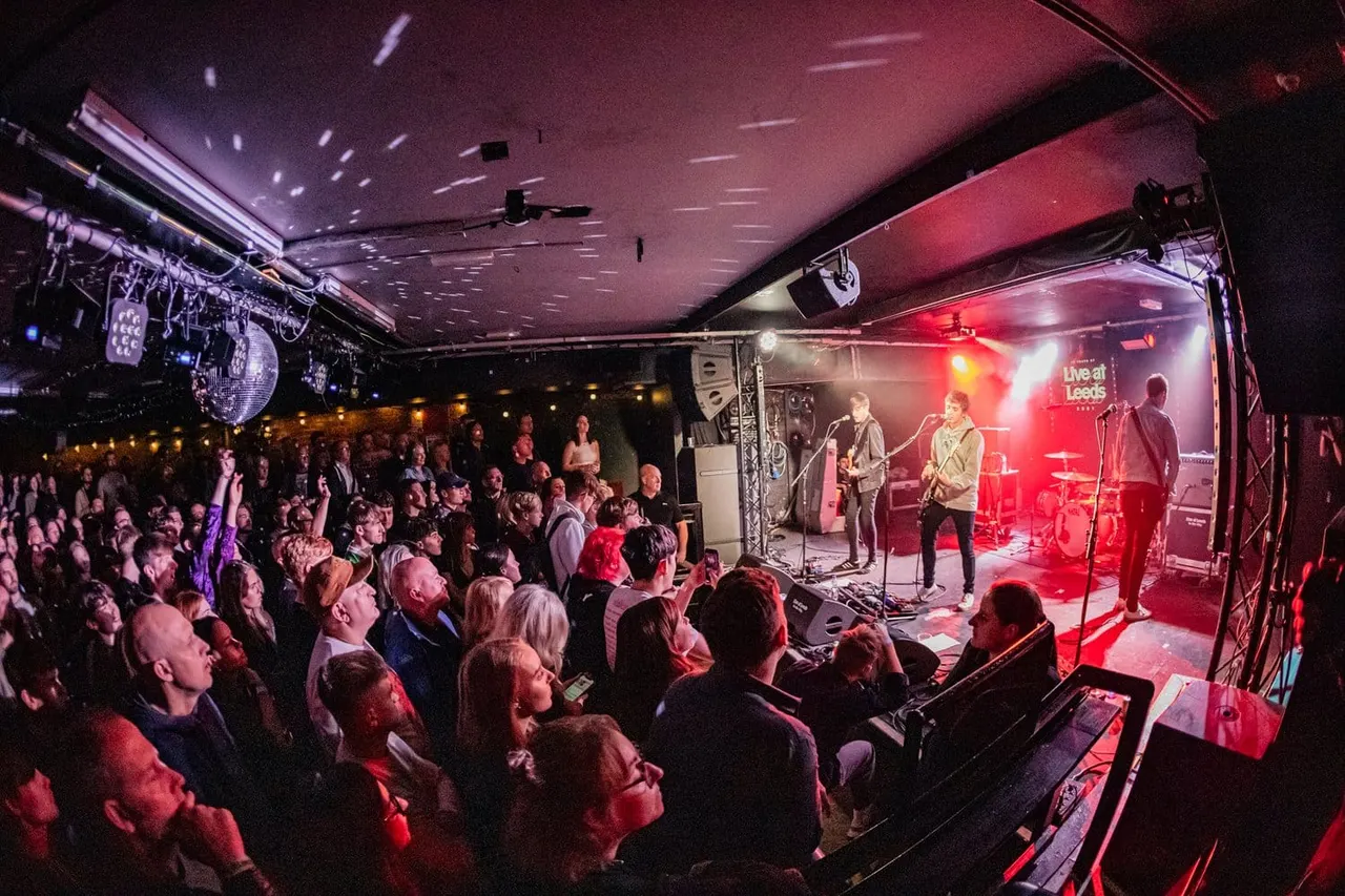 A band performs on an illuminated stage with red lighting while a large crowd of audience members watches from the floor of an indoor music venue.