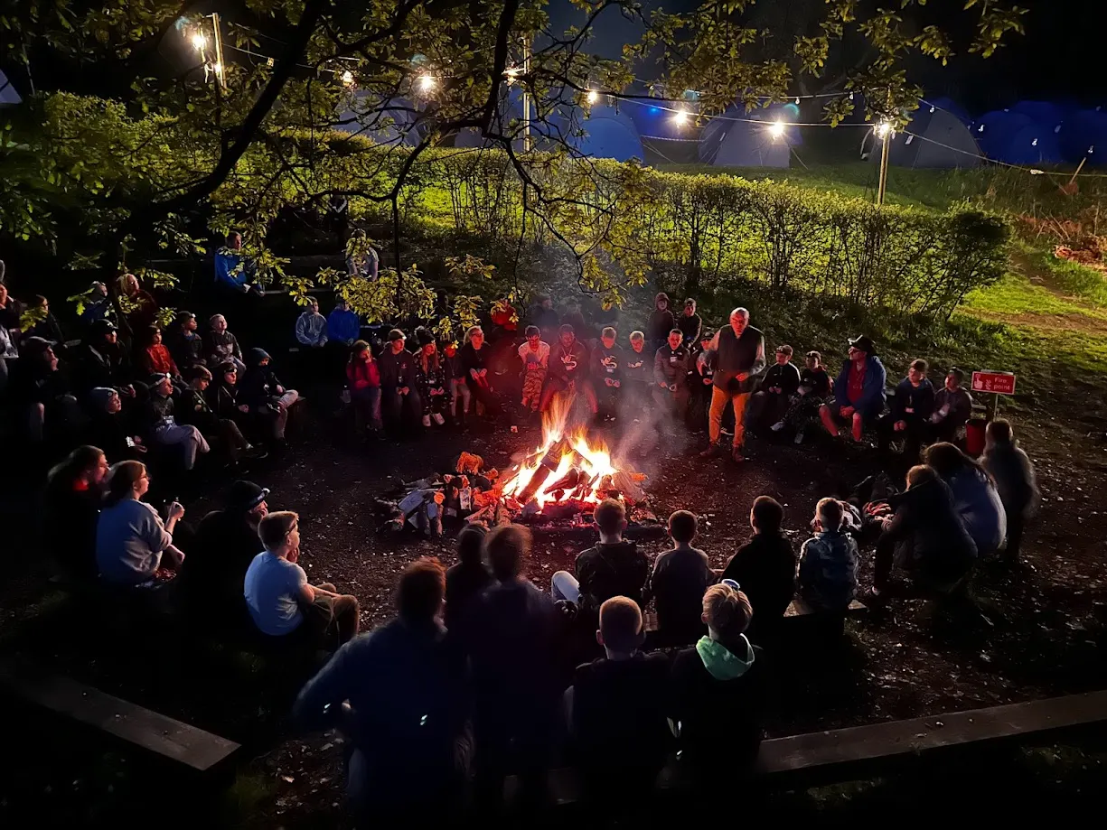 Large group gathered around a blazing bonfire at night under trees with string lights overhead.