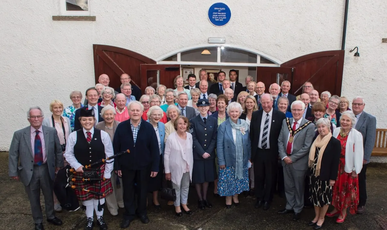 A large group of predominantly older adults posing together outside a white building with red doors, some wearing formal attire and traditional Scottish dress.