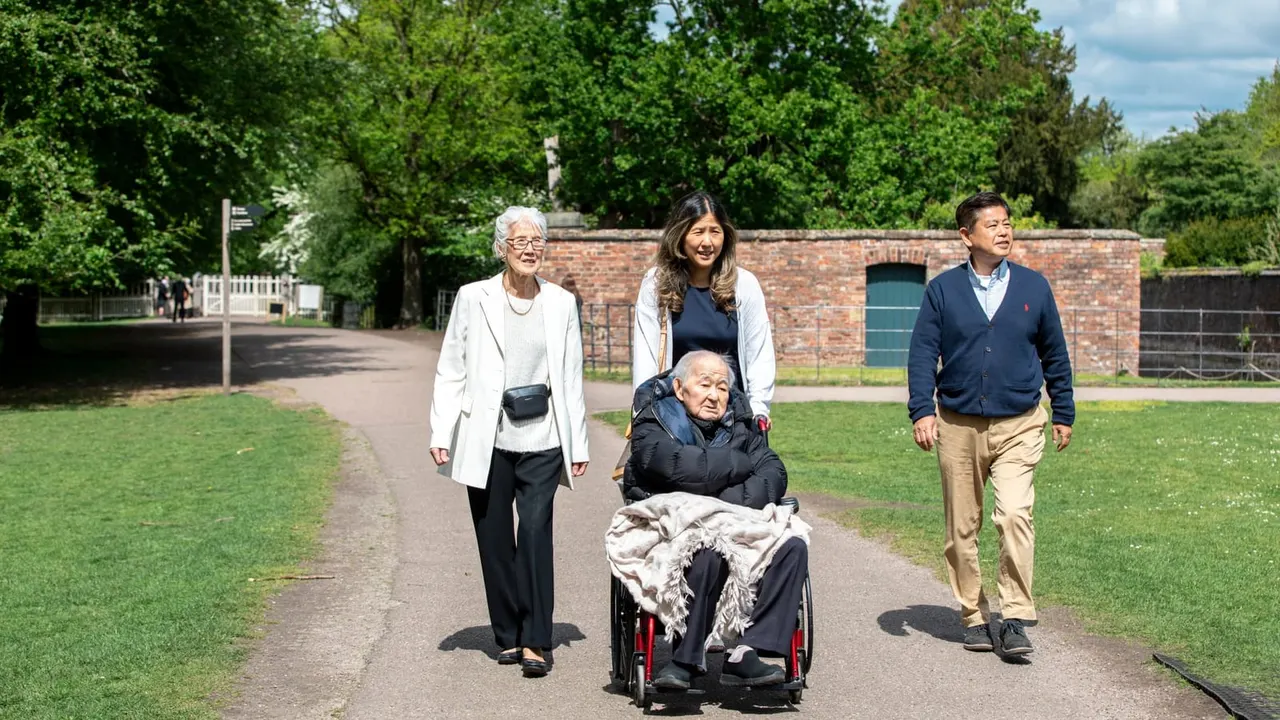 Four people walking along a tree-lined path through a park, one in a wheelchair, with a brick building visible behind them.