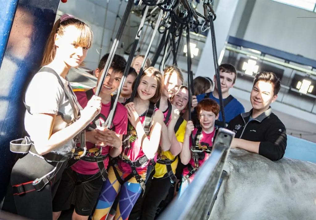 Group of children in harnesses being instructed by an adult at an indoor climbing wall facility.