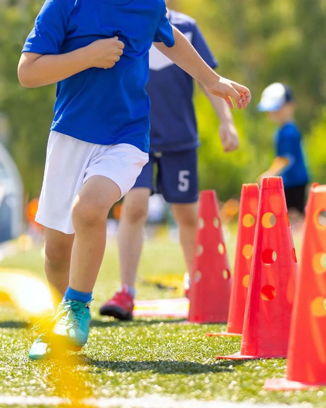 Child in blue shirt running through obstacle course with red cones on grass.