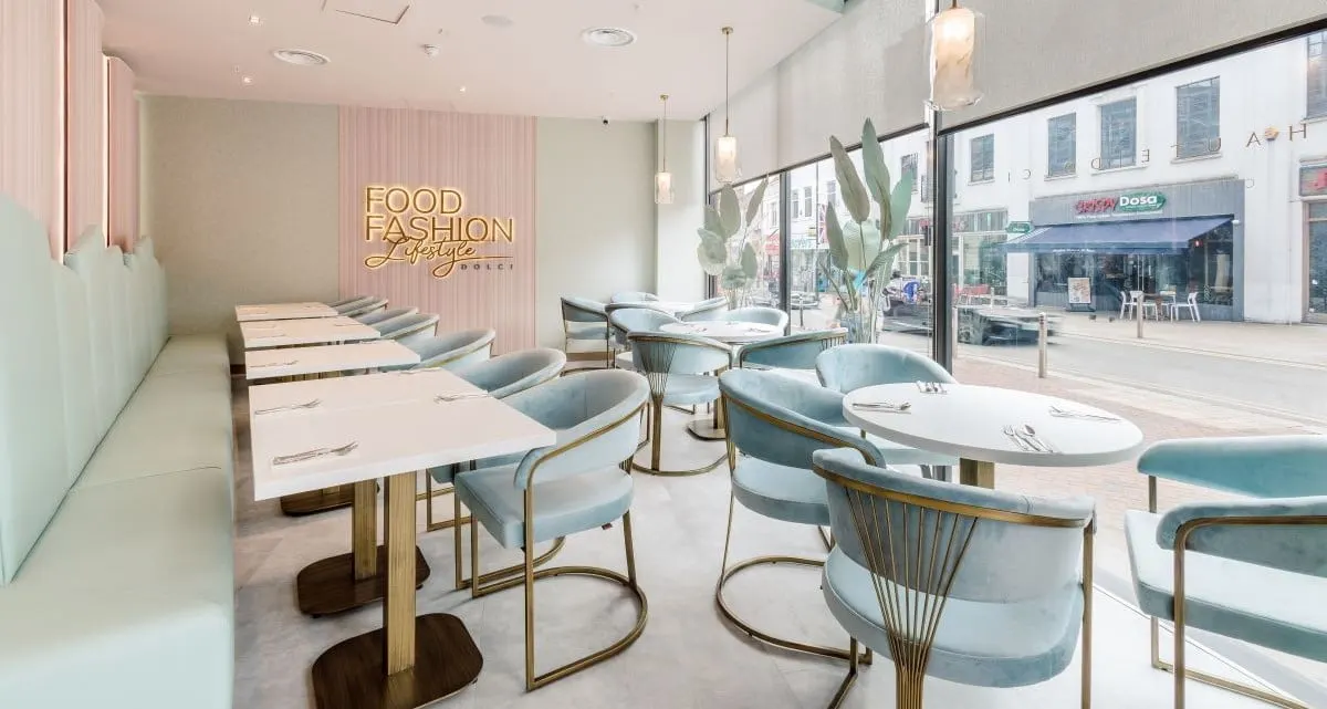 A modern restaurant interior with white tables, teal upholstered chairs, pendant lighting and a neon "Food Fashion Lifestyle" sign visible through large street-facing windows.