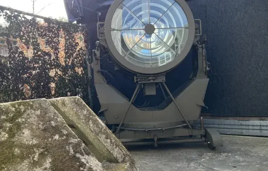 Large vintage aircraft searchlight with illuminated circular lens mounted on metal stand indoors.