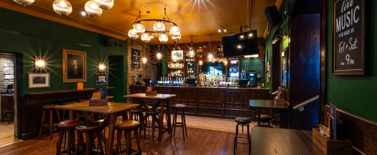 Interior of a pub with wooden bar, high tables and stools, green walls, hanging lights and framed artwork.
