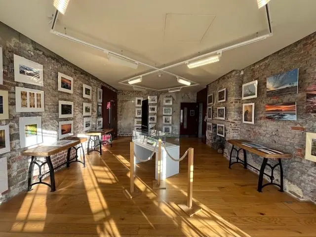 Interior of an art gallery with framed photographs and artworks displayed on exposed brick walls, wooden floors, and display tables.