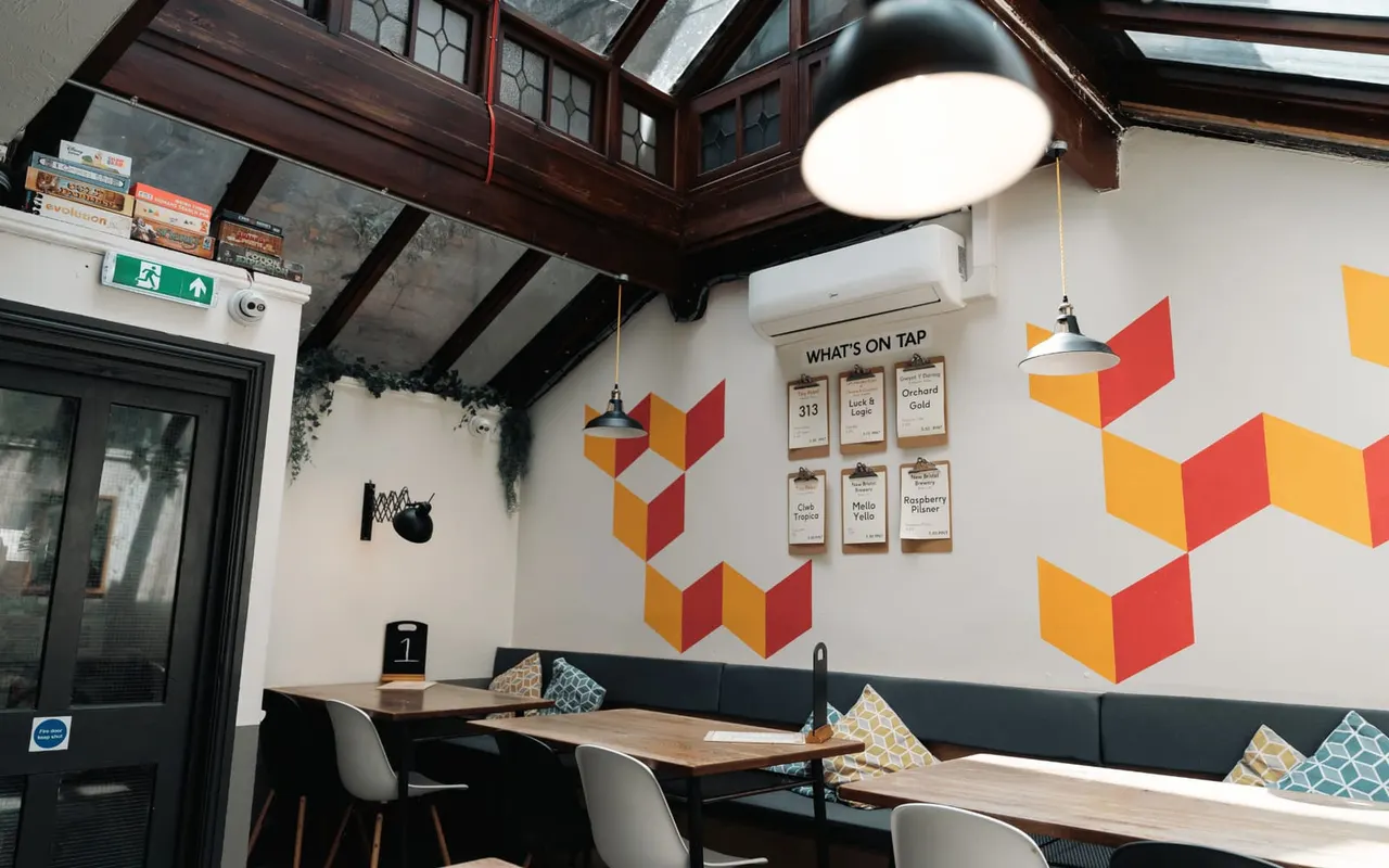 Interior of a board game café with wooden tables, seating booths, and colourful geometric wall art featuring beer tap menus.