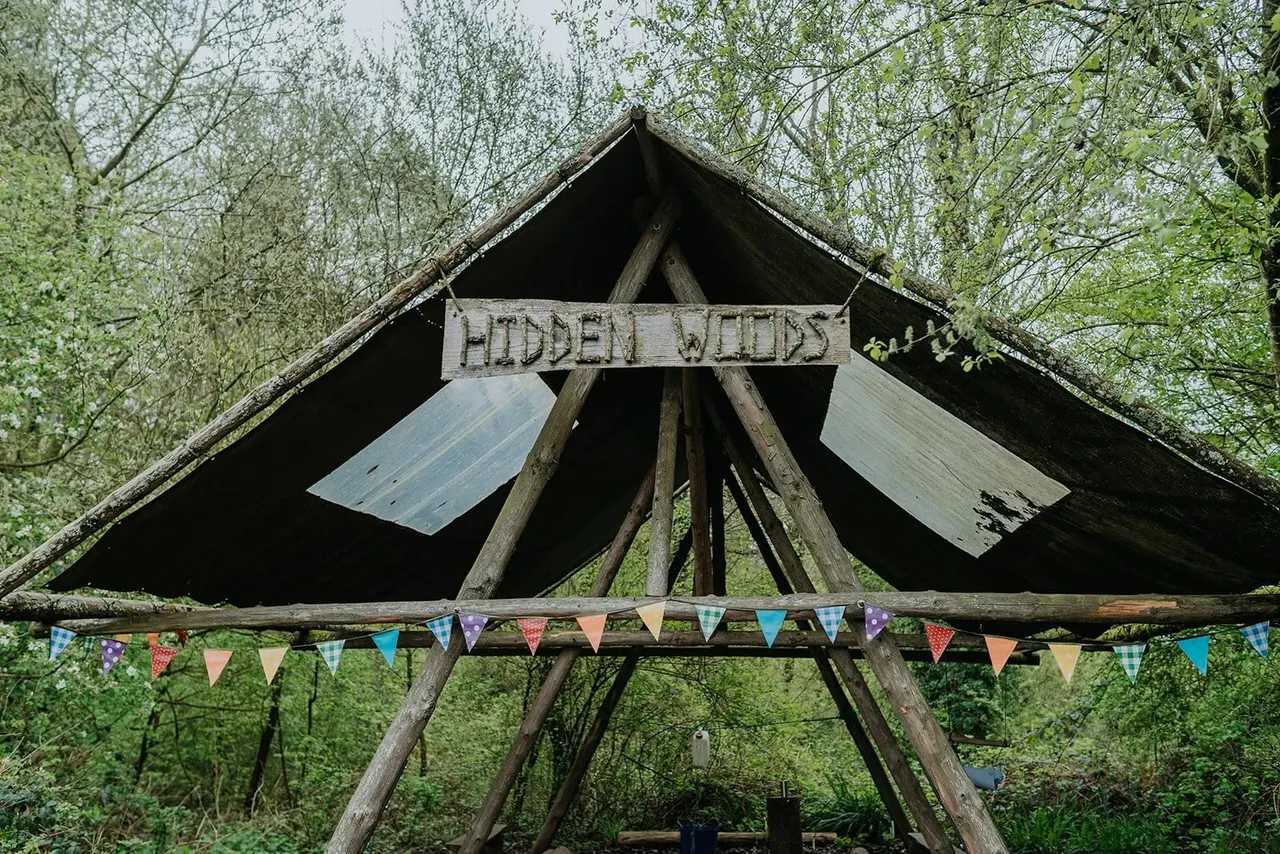 Wooden tipi structure with "Hidden Woods" sign surrounded by green trees and decorated with colourful bunting.