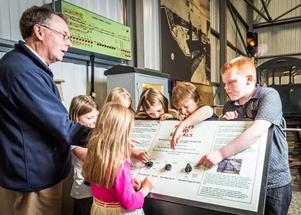 An adult man and five children gather around a large interactive display board inside a railway museum building.