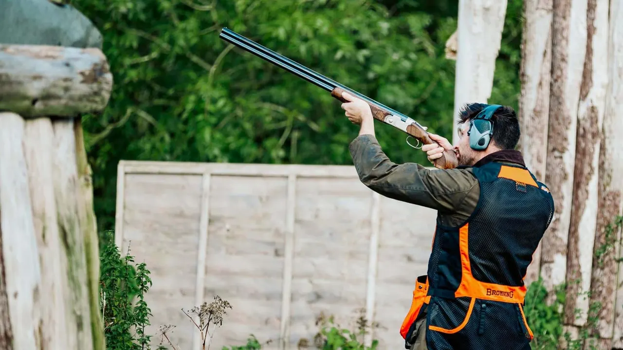 Man in ear defenders and orange safety vest aiming shotgun at clay shooting range with wooden barriers and greenery.