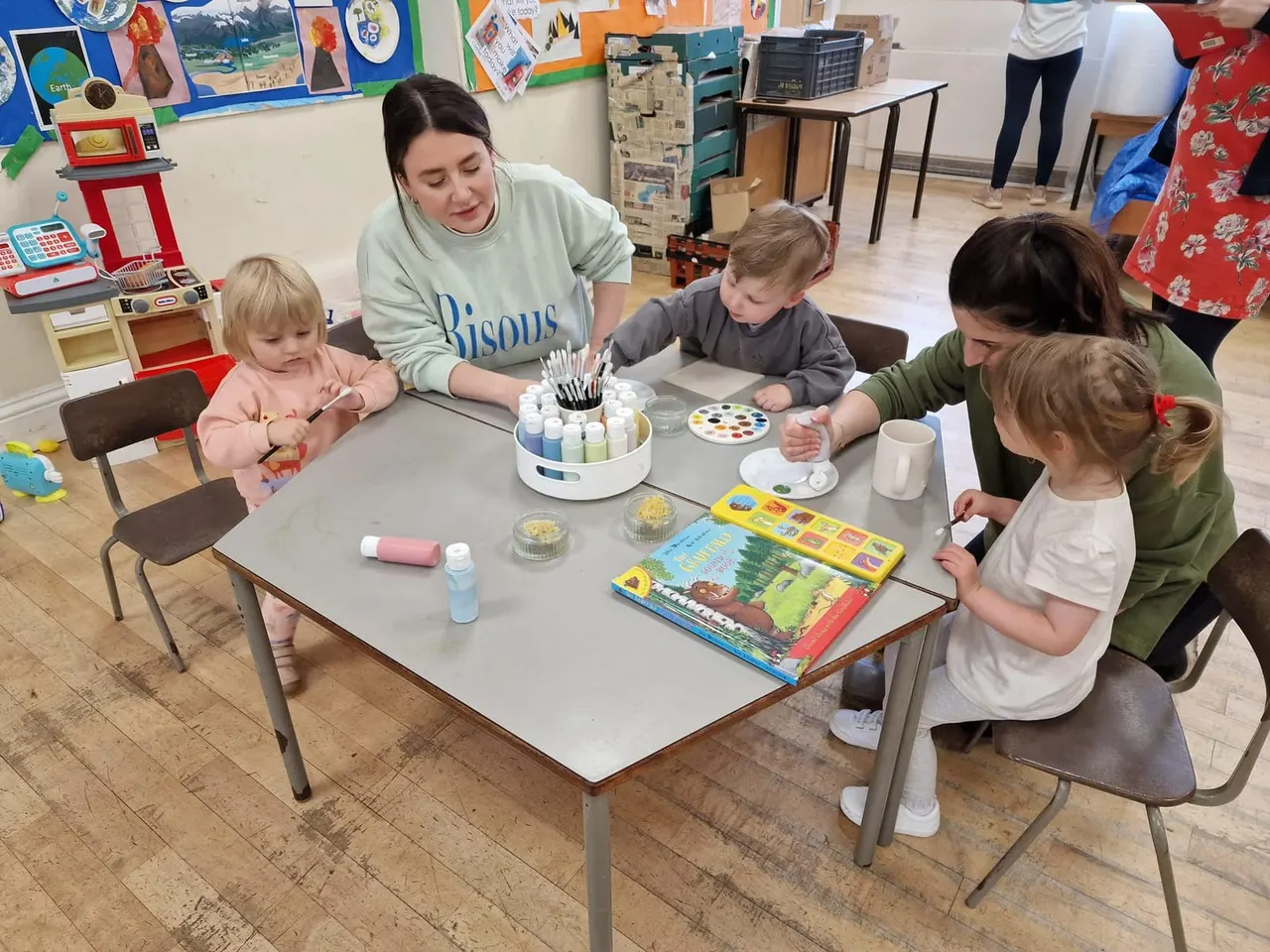 An adult facilitator guides young children in a creative art activity at a table with paints, brushes and colouring materials in an indoor play space.