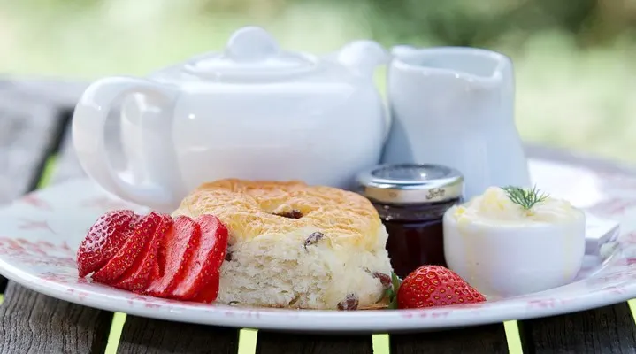 A scone with jam and clotted cream served with fresh strawberries, alongside a teapot and milk jug.
