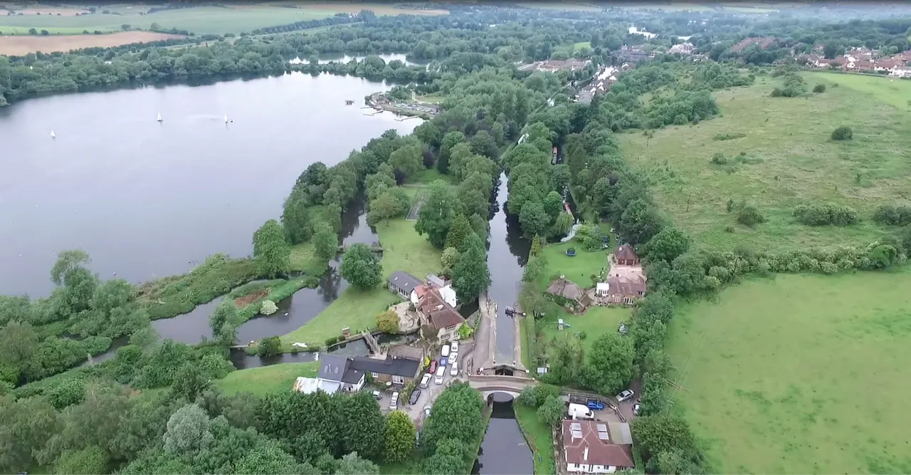 Aerial view of a river valley with locks, tree-lined waterways, green fields, period buildings and a large lake with sailing boats in the distance.