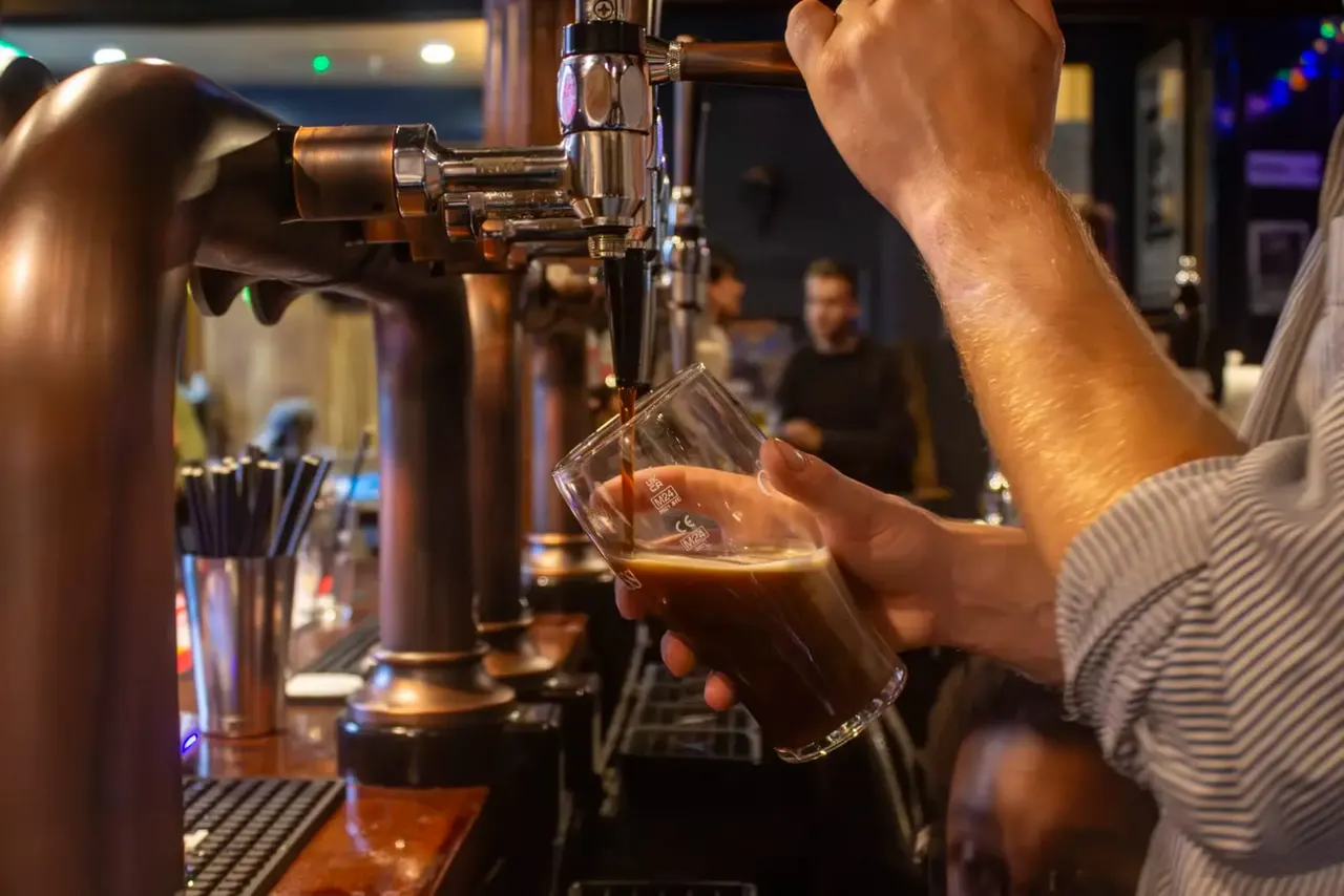Person pouring a pint of beer from a tap at a bar counter with patrons in the background.