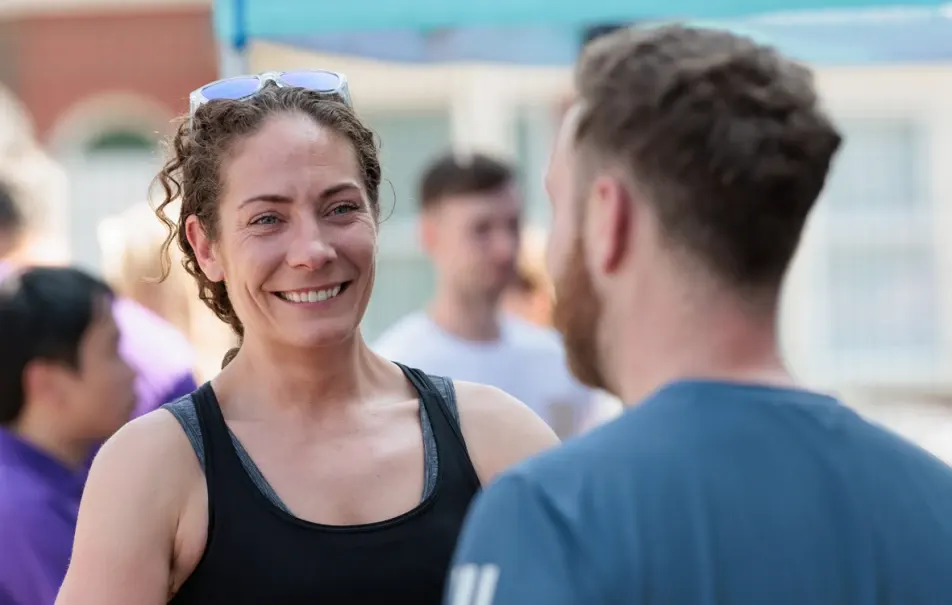 Woman in black tank top smiling whilst talking to a man at an outdoor leisure venue with other people in the background.