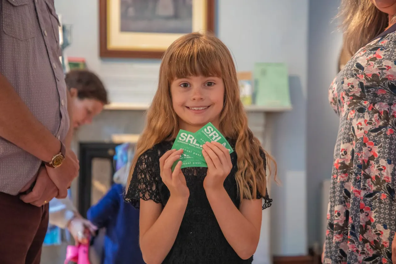 A young girl holding a green activity pass smiles at the camera whilst standing between two adults in an indoor museum space.