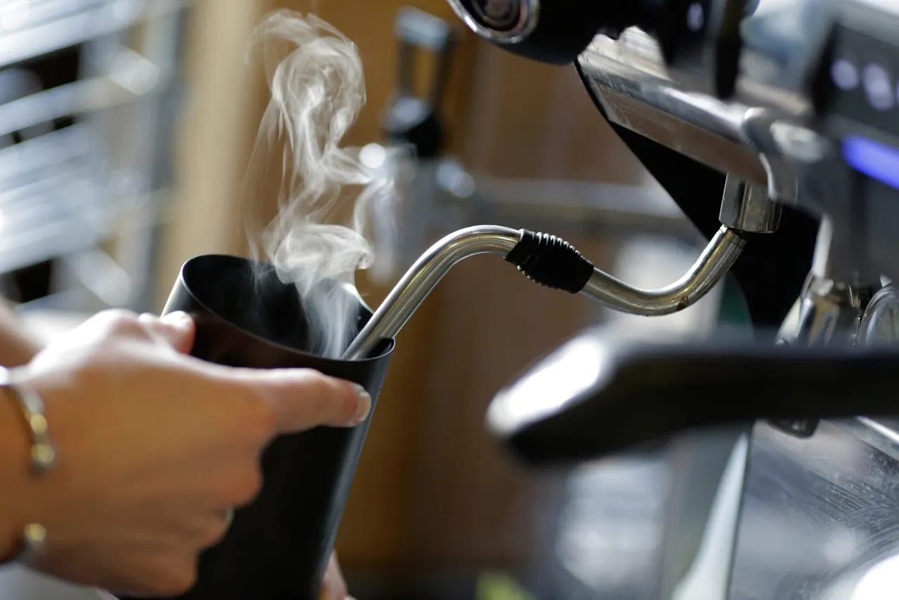 A hand holds a black mug beneath a metal tap dispensing hot steam in an outdoor facility.