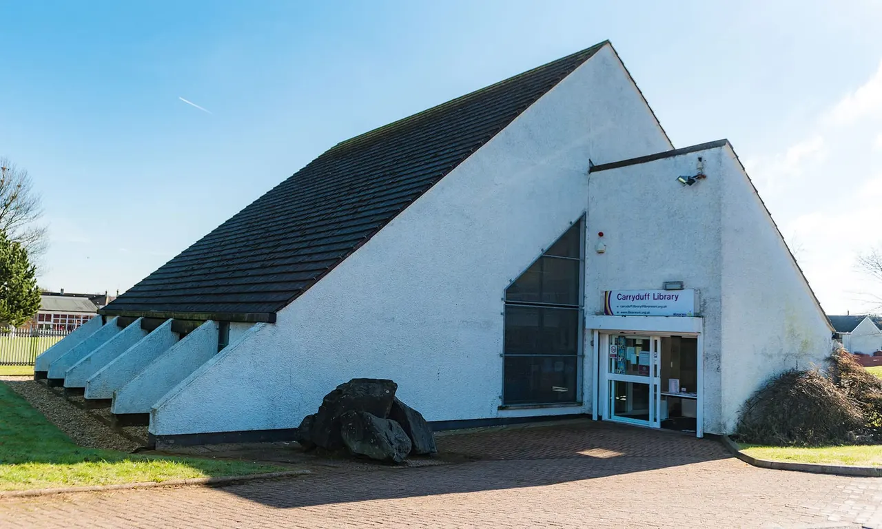 A white-rendered building with a dark slate roof and blue entrance, showing the exterior of Carryduff Library.