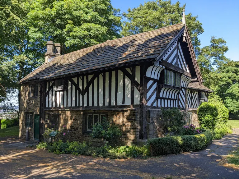 Historic timber-framed house with brick base and decorative black and white half-timbering, surrounded by green trees and landscaping.