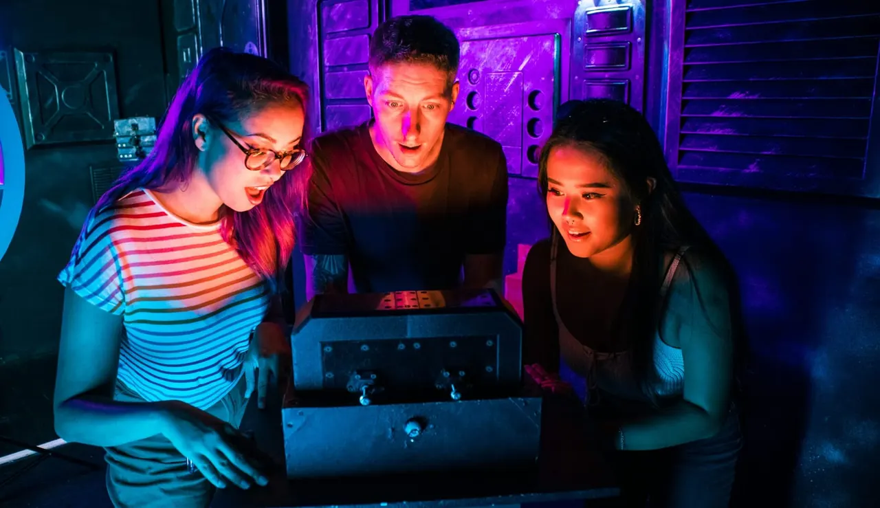 Three players engaged in an escape room puzzle, illuminated by neon purple and cyan lighting with gaming equipment visible.