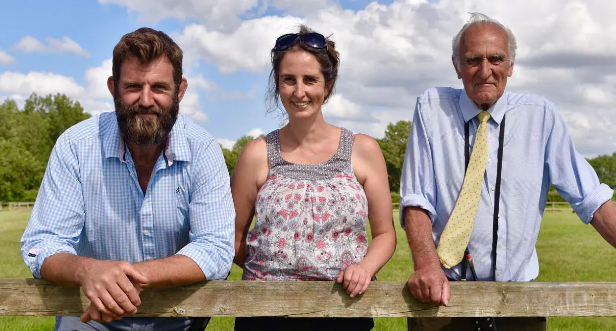 Three people leaning on a wooden fence in a rural field with trees and blue sky behind them.