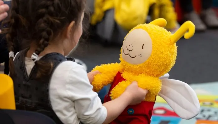 Child holding a yellow soft toy with a smiling face whilst sitting at a table.
