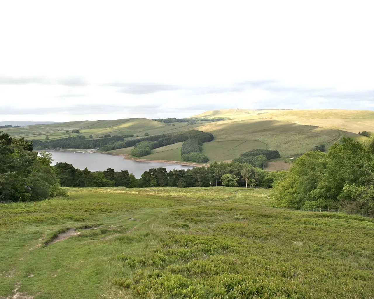 Expansive moorland landscape with a reservoir nestled between forested hills and rolling agricultural fields under cloudy sky.