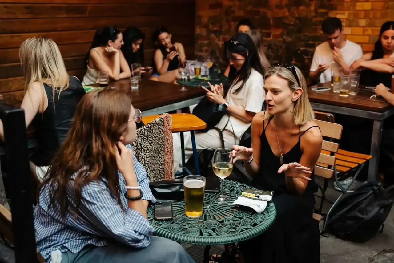 People chatting and drinking at tables and a bar in a brick-walled pub interior.