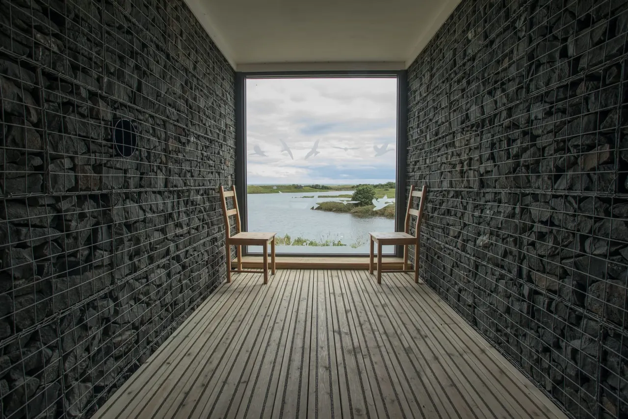 Interior viewing chamber with wooden chairs framing a wide window overlooking wetland landscape with water, islands and cloudy sky.