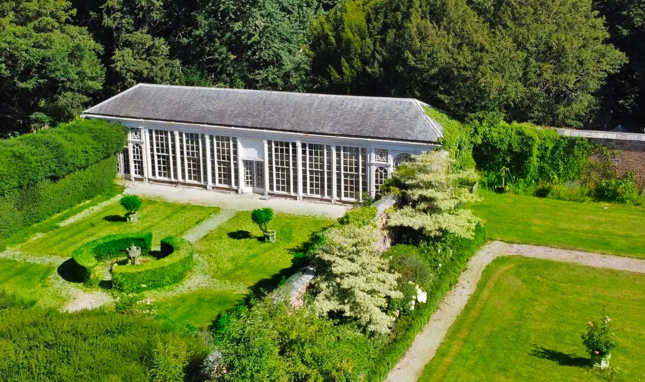Aerial view of a slate-roofed glasshouse surrounded by manicured lawns, topiary, and woodland within a country estate garden.