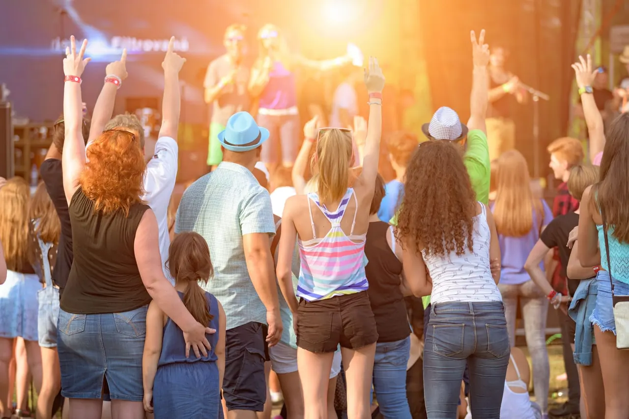 Crowd of people dancing and raising their hands at an outdoor evening event with a stage and performers in warm lighting.