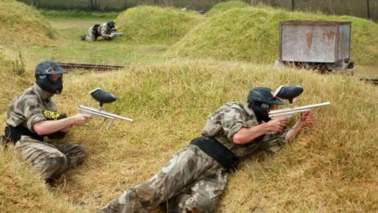 Two paintball players in camouflage gear and masks aim paintball markers whilst crouching on a grassy field.