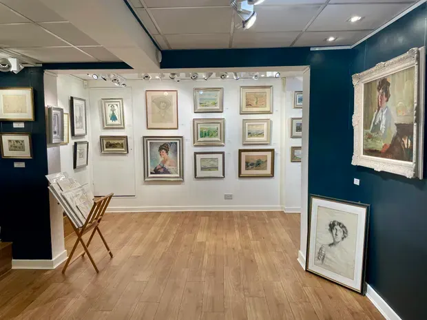 Gallery interior with framed artworks displayed on white and dark teal walls, wooden flooring, and a folding easel.