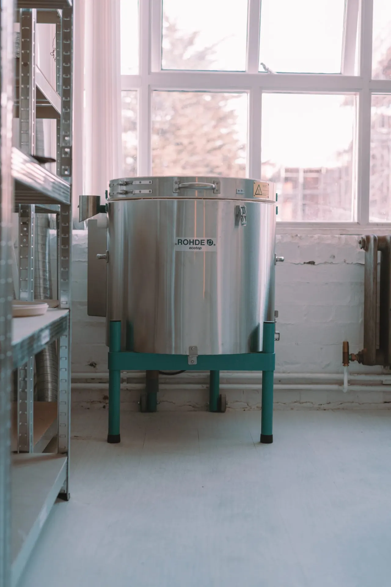 Large stainless steel industrial mixing vessel on green legs in a bright workshop with tall windows overlooking a city.