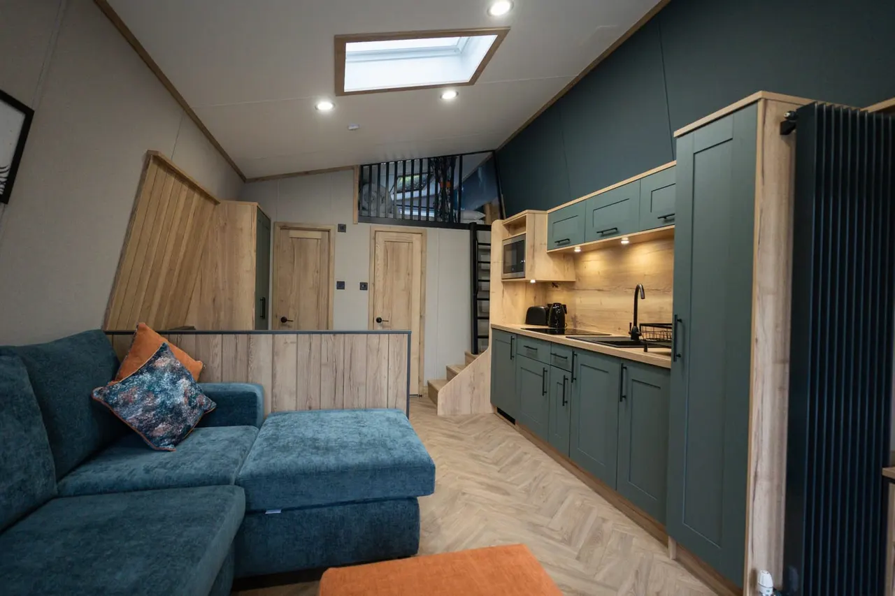 Interior of a modern holiday lodge with a teal and wood colour scheme, featuring a blue sofa, kitchen area, and skylight.